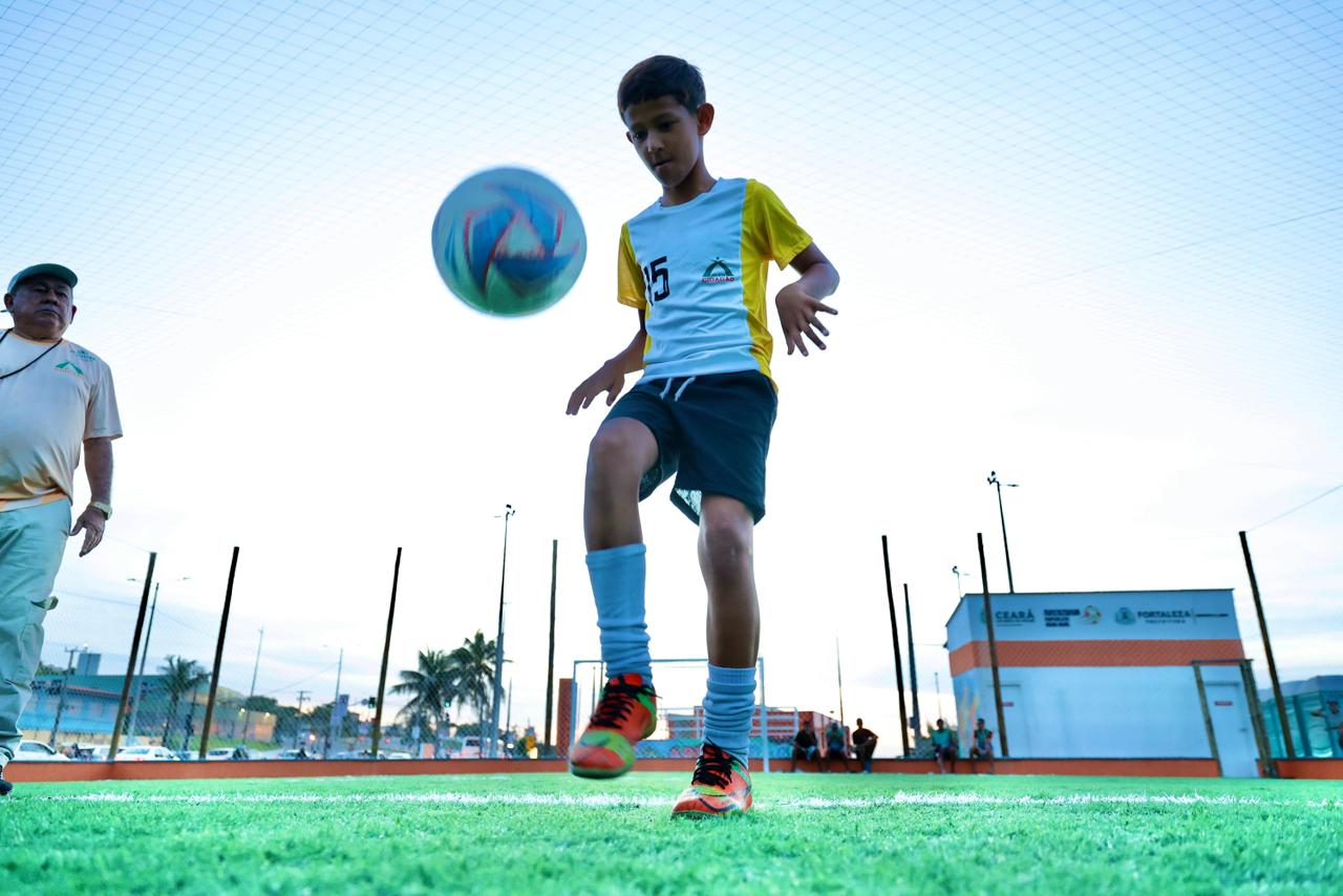 A foto mostra um menino chutando uma bola para o alto, fazendo uma embaixadinha, na areninha requalificada